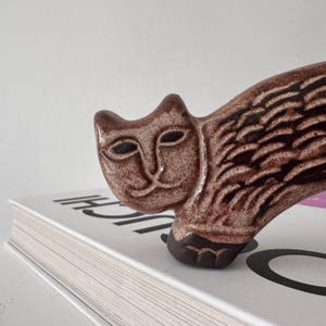 May include: A brown ceramic cat figurine with a stylized design. The cat has a smooth, glossy finish and is positioned on top of a book. The cat's body features a textured pattern, and its face has simple, defined features.