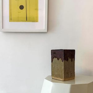 Può includere: Un vaso rettangolare in ceramica con superficie strutturata. La parte superiore del vaso è marrone scuro, con un colore verde e marrone più chiaro nella parte inferiore. Il vaso è esposto su un tavolo bianco, con una stampa gialla incorniciata sullo sfondo.