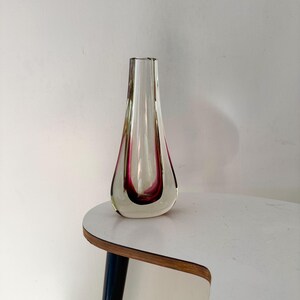 Könnte beinhalten: Eine tropfenförmige Glasvase mit einer klaren äußeren Schicht und einem tief magentafarbenen inneren Kern. Die Vase hat einen schmalen Hals, der sich zu einer abgerundeten Basis hin verbreitert, und besticht durch eine glatte, polierte Oberfläche sowie einen deutlichen Farbkontrast zwischen dem klaren Glas und dem leuchtenden magentafarbenen Zentrum.