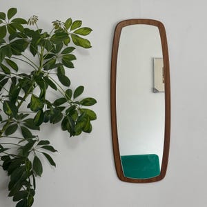 May include: A rectangular mirror with a wooden frame, featuring a rounded top and bottom. The mirror has a green accent at the base. A leafy green plant is visible on the left. The mirror's dimensions are approximately 100 cm in height.