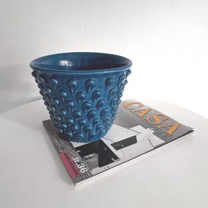 Op de afbeelding: Een teal keramische plantenbak met een textuur van druppelvormige patronen. De plantenbak staat op een tijdschrift met het woord "CASA" zichtbaar. De plantenbak heeft een brede opening en een afgeronde basis.