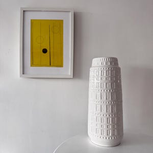 Peut inclure: Un vase en céramique blanc avec un motif géométrique texturé est posé sur une surface blanche. Une œuvre d'art jaune encadrée avec des lignes et des cercles noirs est accrochée au mur blanc derrière le vase.