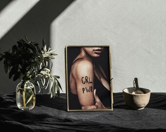 May include: A framed print with the text "CRL PWR" on a person's arm. The image is in a gold frame, and the background includes a vase with greenery and a small bowl with a spoon. The scene is set on a dark surface.