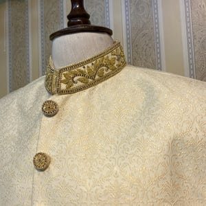 May include: Cream-coloured formal jacket with elaborate gold embroidery on the collar and decorative buttons. The jacket has a subtle, repeating pattern across the fabric. The design suggests a formal or ceremonial garment.