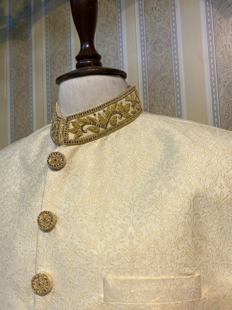 May include: Cream-coloured formal jacket with elaborate gold embroidery on the collar and decorative buttons. The jacket has a subtle, repeating pattern across the fabric. The design suggests a formal or ceremonial garment.
