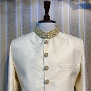 May include: Ivory formal jacket with a mandarin collar and gold embroidery. The jacket has gold buttons and a subtle floral pattern. The sleeves have gold embroidery at the cuffs. The jacket is displayed on a mannequin.
