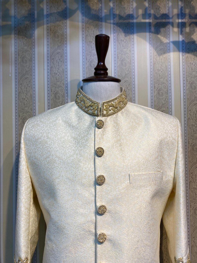 May include: Ivory formal jacket with a mandarin collar and gold embroidery. The jacket has gold buttons and a subtle floral pattern. The sleeves have gold embroidery at the cuffs. The jacket is displayed on a mannequin.