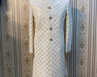 Könnte beinhalten: Cremefarbene, langärmlige Jacke mit Stehkragen und geometrischem Muster. Die Jacke hat goldfarbene Akzente an Kragen, Manschetten und Knöpfen. Das Kleidungsstück wird auf einer Schaufensterpuppe präsentiert.