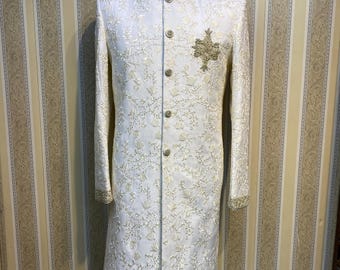 Könnte beinhalten: Eine cremefarbene Sherwani mit langen Ärmeln und aufwendiger goldener Stickerei. Die formelle Jacke hat einen Stehkragen, dekorative Knöpfe und ein goldgesticktes Emblem auf der Brust. Die Ärmel und der Kragen sind mit Gold verziert.