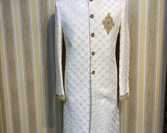 May include: A cream-coloured, long-sleeved formal jacket with a mandarin collar. The jacket features a diamond pattern with gold embroidery, gold buttons, and gold trim on the collar and cuffs. A gold embroidered emblem is on the chest.