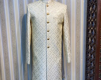 May include: A cream-coloured, long-sleeved jacket with a geometric pattern and gold-coloured buttons. The jacket is displayed on a mannequin. The jacket has a mandarin collar and a tailored fit, suitable for formal occasions.
