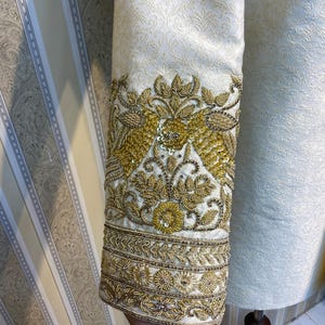 May include: Close-up of a cream-coloured garment sleeve with intricate gold embroidery. The sleeve features a floral pattern and detailed embellishments. The design includes gold sequins and beads, creating a luxurious and ornate appearance.