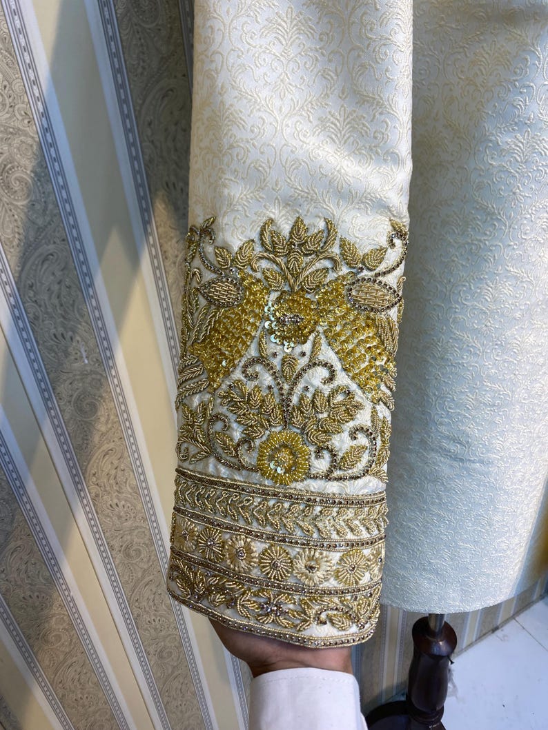 May include: Close-up of a cream-coloured garment sleeve with intricate gold embroidery. The sleeve features a floral pattern and detailed embellishments. The design includes gold sequins and beads, creating a luxurious and ornate appearance.