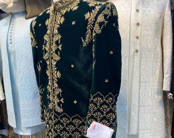 May include: A dark green velvet jacket with detailed gold embroidery. The jacket has a high collar and elaborate floral and geometric designs. Other garments in white and gold are visible in the background, suggesting formal attire.