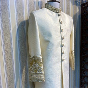 May include: Cream-coloured formal jacket with gold embroidery on the collar and cuffs. The jacket has a mandarin collar and decorative gold buttons. The fabric has a subtle, textured pattern, and the sleeves are long.