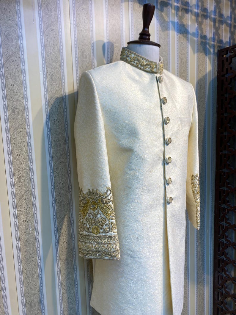 May include: Cream-coloured formal jacket with gold embroidery on the collar and cuffs. The jacket has a mandarin collar and decorative gold buttons. The fabric has a subtle, textured pattern, and the sleeves are long.