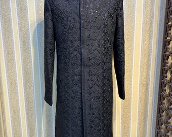 Könnte beinhalten: Schwarze, langärmlige formelle Jacke mit aufwendiger floraler Stickerei. Die Jacke hat einen Stehkragen und eine Knopfleiste vorne. Das Kleidungsstück wird auf einer Schaufensterpuppe präsentiert, die die maßgeschneiderte Passform und das detaillierte Design zeigt. Die Jacke reicht bis zur Mitte des Oberschenkels.
