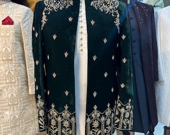 May include: A dark green velvet jacket with gold embroidery on the collar, shoulders, and cuffs. The jacket is paired with a white kurta. Other garments are visible in the background, including a burgundy turban and a cream-coloured jacket.