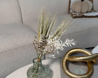 May include: A clear glass vase with a textured design holds a bouquet of dried flowers and greenery. A gold-colored decorative knot and a book are on a white table. A beige couch and a pumpkin-shaped pillow are in the background.