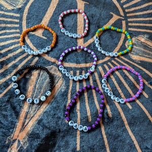 Op de afbeelding: Een verzameling kralenarmbanden in verschillende kleuren, elk met een woord met witte letterkralen. De armbanden zijn oranje, roze, geel, blauw, paars en zwart. De woorden zijn "MELIORA", "COPIA", "MOON", "FUCKING", "PAPA", "PHANTOM" en "PETUA".