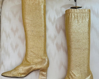 May include: A pair of knee-high boots in a metallic gold finish. These boots feature a block heel, a rounded toe, and a gathered elasticized top edge. Each boot is fitted with a dark brown plastic shoe tree insert for shape retention.