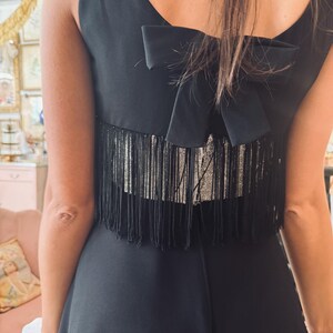 Pode incluir: Vestido preto sem mangas com um la&ccedil;o nas costas. O vestido apresenta um detalhe de franjas nas costas. O vestido &eacute; de cor preta lisa e a franja parece ser prateada.