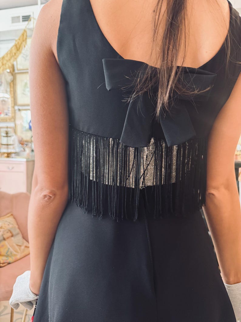 Pode incluir: Vestido preto sem mangas com um la&ccedil;o nas costas. O vestido apresenta um detalhe de franjas nas costas. O vestido &eacute; de cor preta lisa e a franja parece ser prateada.
