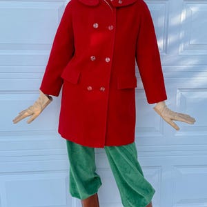 K&ouml;nnte beinhalten: Ein roter Vintage-Doppelreiher-Mantel mit gro&szlig;en Kn&ouml;pfen, kombiniert mit gr&uuml;ner Samthose und braunen Lederstiefeln. Das Outfit umfasst beige Handschuhe und einen Pelzhut mit Sonnenbrille.