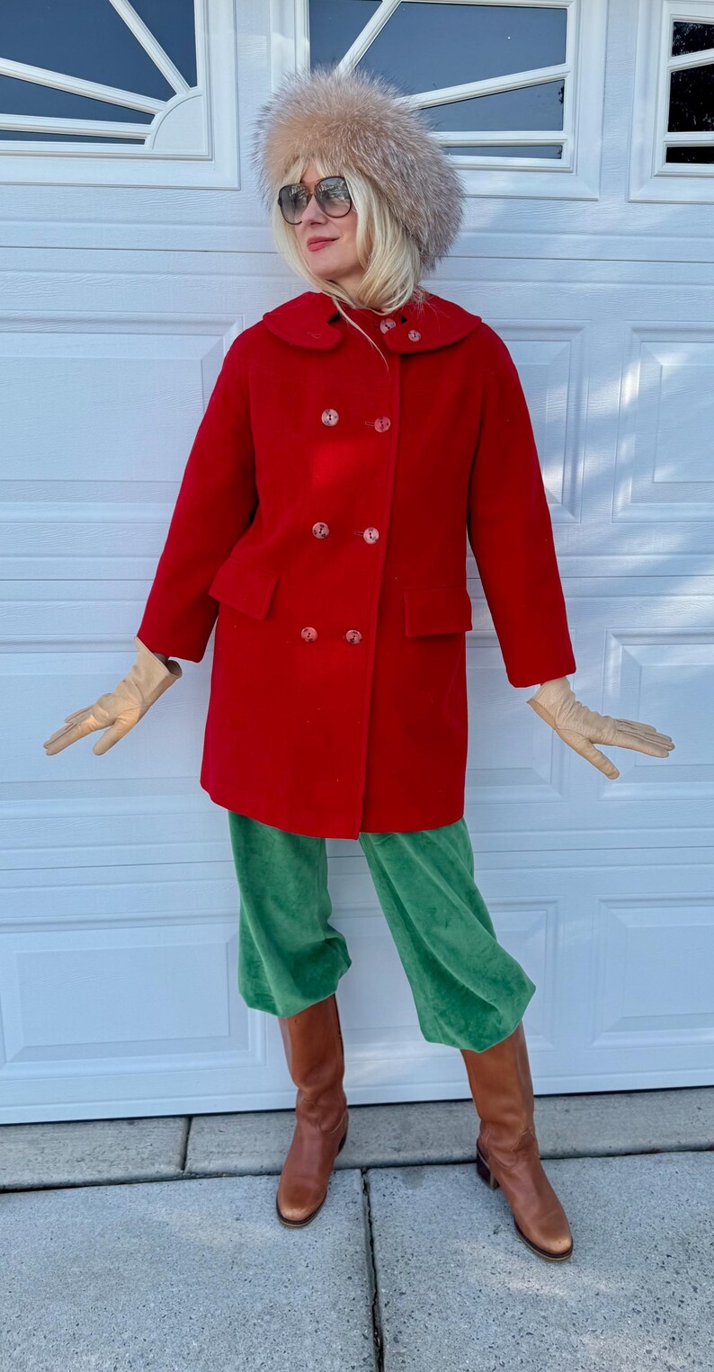 K&ouml;nnte beinhalten: Ein roter Vintage-Doppelreiher-Mantel mit gro&szlig;en Kn&ouml;pfen, kombiniert mit gr&uuml;ner Samthose und braunen Lederstiefeln. Das Outfit umfasst beige Handschuhe und einen Pelzhut mit Sonnenbrille.