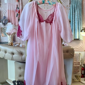 May include: A sheer, light pink vintage nightgown with a matching robe. The nightgown features a deep burgundy lace trim on the neckline, sleeves, and hem. The robe is open-fronted and hangs on a gold clothing rack. Other vintage dresses are visible in the background.