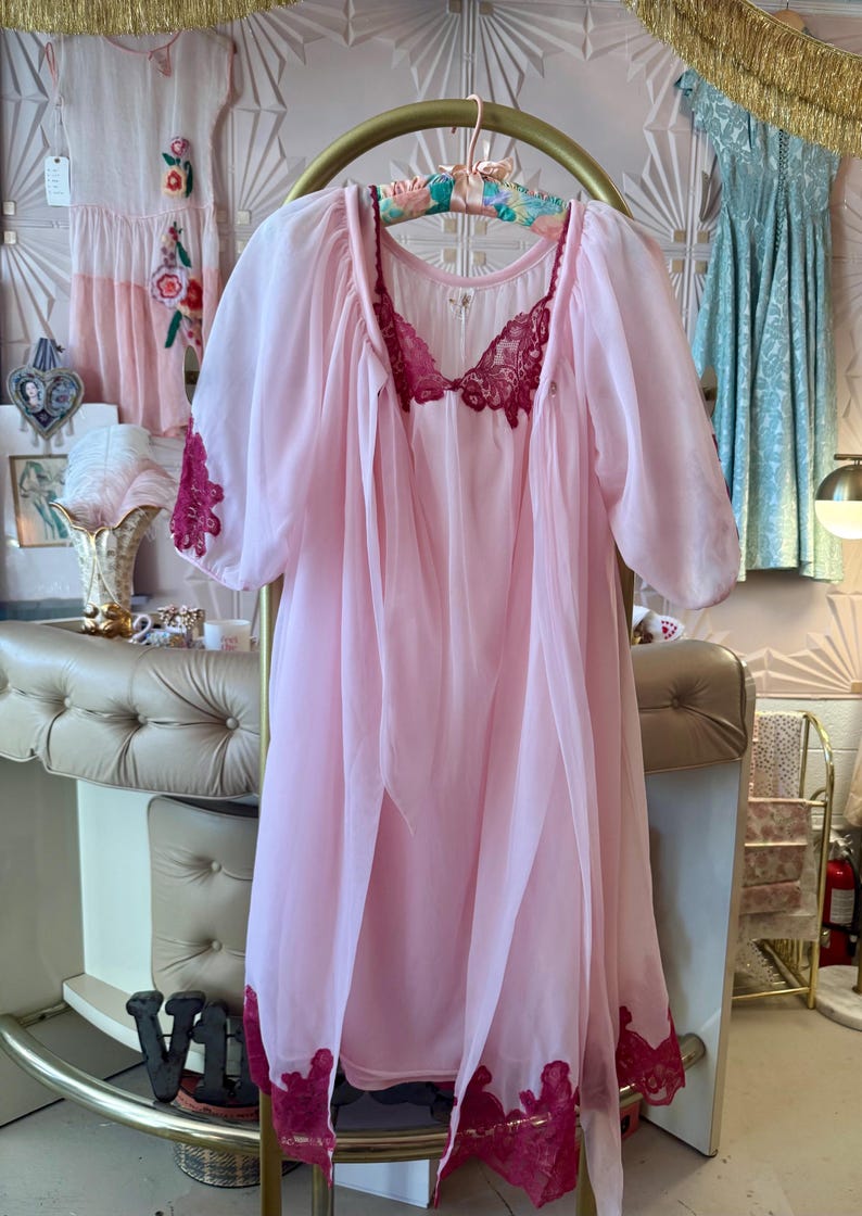 May include: A sheer, light pink vintage nightgown with a matching robe. The nightgown features a deep burgundy lace trim on the neckline, sleeves, and hem. The robe is open-fronted and hangs on a gold clothing rack. Other vintage dresses are visible in the background.
