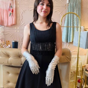 Pode incluir: Um vestido preto com franjas na cintura e luvas prateadas. O vestido tem um design sem mangas e uma saia rodada. As luvas s&atilde;o longas e feitas de um material brilhante. O fundo inclui outros vestidos e elementos decorativos.