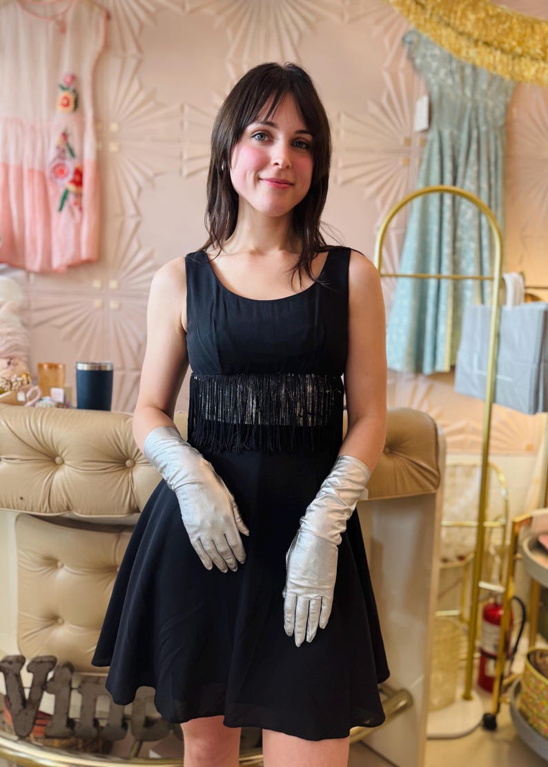 Pode incluir: Um vestido preto com franjas na cintura e luvas prateadas. O vestido tem um design sem mangas e uma saia rodada. As luvas s&atilde;o longas e feitas de um material brilhante. O fundo inclui outros vestidos e elementos decorativos.