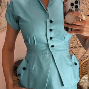 Pode incluir: Um fato azul claro de estilo vintage. A parte de cima tem gola, mangas curtas e fecho de bot&otilde;es. O fato inclui uma saia a condizer, e a parte de cima tem cintura justa e bolsos decorativos. O fato &eacute; feito de tecido liso.