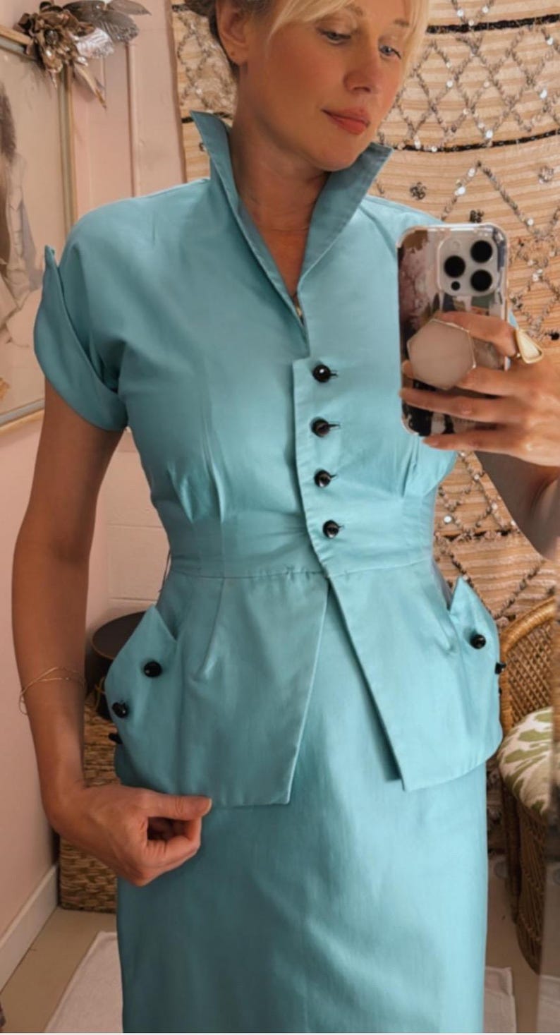 Pode incluir: Um fato azul claro de estilo vintage. A parte de cima tem gola, mangas curtas e fecho de bot&otilde;es. O fato inclui uma saia a condizer, e a parte de cima tem cintura justa e bolsos decorativos. O fato &eacute; feito de tecido liso.