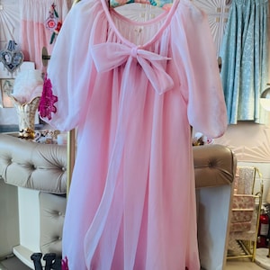May include: A sheer, light pink vintage robe with a large bow at the neckline and a dark pink lace trim along the hem and sleeves. The robe is hanging on a floral print hanger.