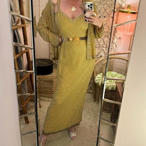 Può includere: Uno specchio a figura intera riflette una donna che indossa un abito dorato con paillettes, un cardigan abbinato e una cintura dorata. L'abito ha spalline sottili e scollo a V. Indossa anche tacchi bianchi. Lo sfondo include una sedia in vimini e un arazzo decorativo.