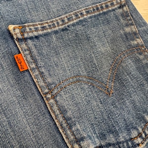 May include: Close-up of a blue denim jeans pocket with brown stitching. An orange tag with the word "LEVI'S" is attached to the pocket. The pocket has a curved design and is sewn onto the jeans.