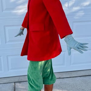 K&ouml;nnte beinhalten: Ein leuchtend roter Mantel mit einer einzelnen Tasche, kombiniert mit gr&uuml;ner Samthose und braunen Lederstiefeln. Das Outfit wird durch hellblaue Handschuhe erg&auml;nzt.