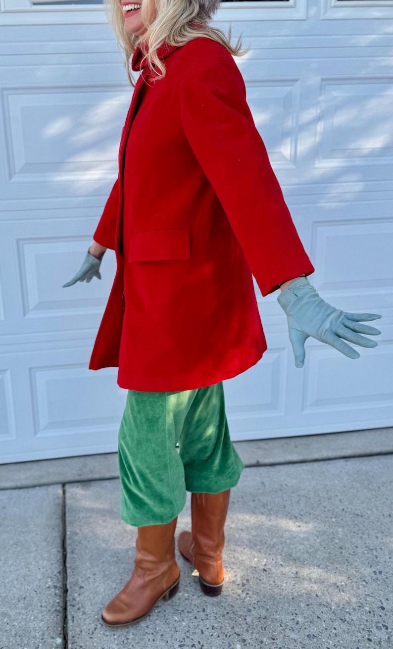 K&ouml;nnte beinhalten: Ein leuchtend roter Mantel mit einer einzelnen Tasche, kombiniert mit gr&uuml;ner Samthose und braunen Lederstiefeln. Das Outfit wird durch hellblaue Handschuhe erg&auml;nzt.