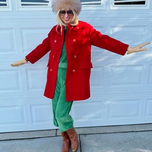 K&ouml;nnte beinhalten: Eine Person tr&auml;gt einen roten Mantel, einen gr&uuml;nen Samt-Jumpsuit, einen Pelzhut, eine Sonnenbrille und braune Stiefel. Der zweireihige Mantel hat gro&szlig;e Kn&ouml;pfe. Das Outfit ist eine Mischung aus Texturen und Farben.