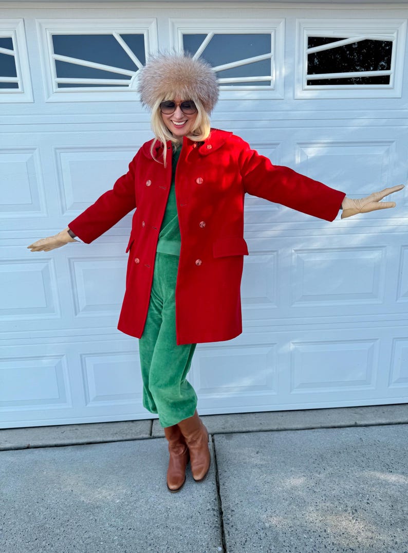 K&ouml;nnte beinhalten: Eine Person tr&auml;gt einen roten Mantel, einen gr&uuml;nen Samt-Jumpsuit, einen Pelzhut, eine Sonnenbrille und braune Stiefel. Der zweireihige Mantel hat gro&szlig;e Kn&ouml;pfe. Das Outfit ist eine Mischung aus Texturen und Farben.