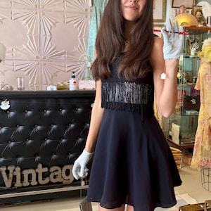 Pode incluir: Um vestido vintage preto com franjas no corpete. O vestido tem uma saia rodada e &eacute; combinado com uma pequena bolsa de contas. A palavra "Vintage" &eacute; vis&iacute;vel em um balc&atilde;o preto.