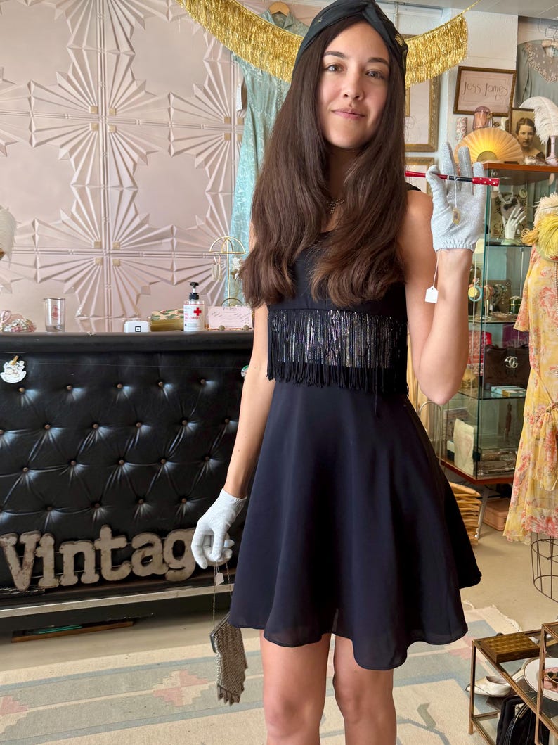 Pode incluir: Um vestido vintage preto com franjas no corpete. O vestido tem uma saia rodada e &eacute; combinado com uma pequena bolsa de contas. A palavra "Vintage" &eacute; vis&iacute;vel em um balc&atilde;o preto.
