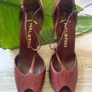 May include: A pair of brown leather Halston high heels with open toes and ankle straps. The heels have a t-strap design and are set against a neutral background. The words "MADE IN ITALY" and "HALSTON" are visible on the insole.