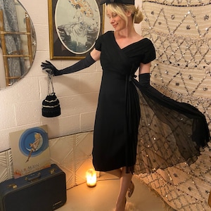Puede incluir: Un vestido negro vintage con una superposición de falda plisada transparente. El vestido tiene mangas cortas y un escote en V. El atuendo se complementa con guantes largos negros, un sombrero negro pequeño y tacones dorados. Un pequeño bolso negro con cuentas cuelga de la mano enguantada.