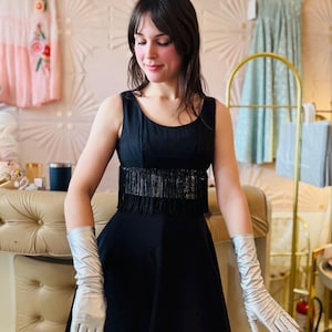 Pode incluir: Um vestido preto sem mangas com franjas na cintura. O vestido &eacute; combinado com luvas longas prateadas. O fundo apresenta um interior em estilo vintage com elementos decorativos e outros vestidos.