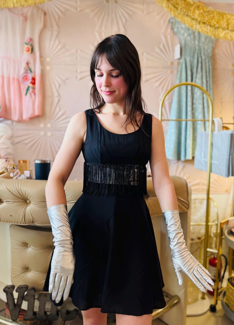 Pode incluir: Um vestido preto sem mangas com franjas na cintura. O vestido &eacute; combinado com luvas longas prateadas. O fundo apresenta um interior em estilo vintage com elementos decorativos e outros vestidos.