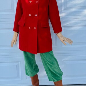 K&ouml;nnte beinhalten: Ein roter, zweireihiger Mantel mit gro&szlig;en Kn&ouml;pfen, kombiniert mit einer gr&uuml;nen Samthose und braunen Stiefeln. Das Outfit wird durch einen Pelzhut und Handschuhe erg&auml;nzt. Der Mantel hat zwei Vordertaschen.