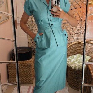 Pode incluir: Um conjunto de duas pe&ccedil;as azul claro em estilo vintage. Inclui um top com gola e mangas curtas com bot&otilde;es pretos e uma saia a condizer at&eacute; ao joelho. O conjunto &eacute; completado com sapatilhas pretas. A est&eacute;tica geral &eacute; retro e elegante.